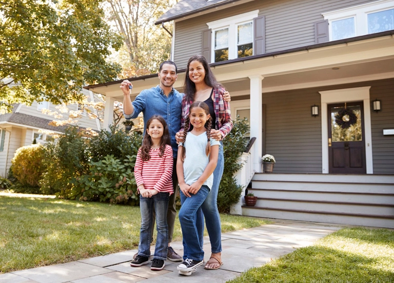 Uma família de quatro pessoas em pé em frente a uma casa suburbana de dois andares.