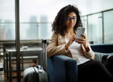 A-woman-scrolls-on-a-cell-phone-while-seated-in-a-blue-velvet-armchair-with-luggage-beside-her.